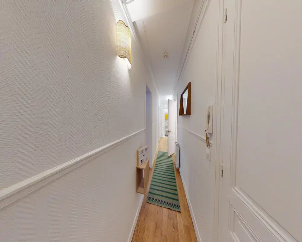 Narrow, well-lit entrance corridor with white textured walls, wooden floor and a green runner rug leading to inner rooms. A wall lamp and mirror add functional decor.