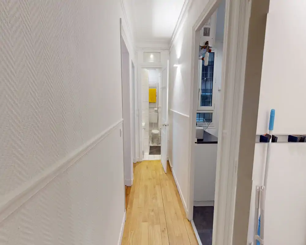 Bright, narrow hallway with light wood flooring leading to a small bathroom at the end; clean white walls and simple modern lighting.