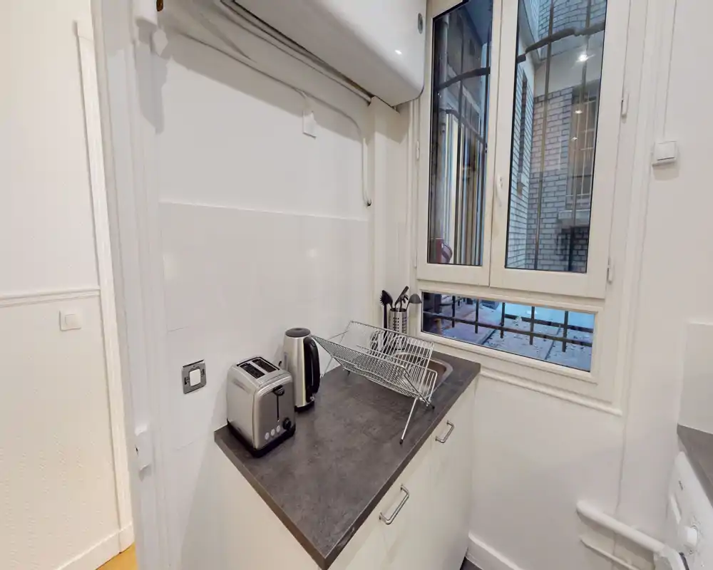Compact modern kitchenette with a dark countertop, toaster, electric kettle and a dish rack beside a window letting in natural light — practical and tidy for short stays.
