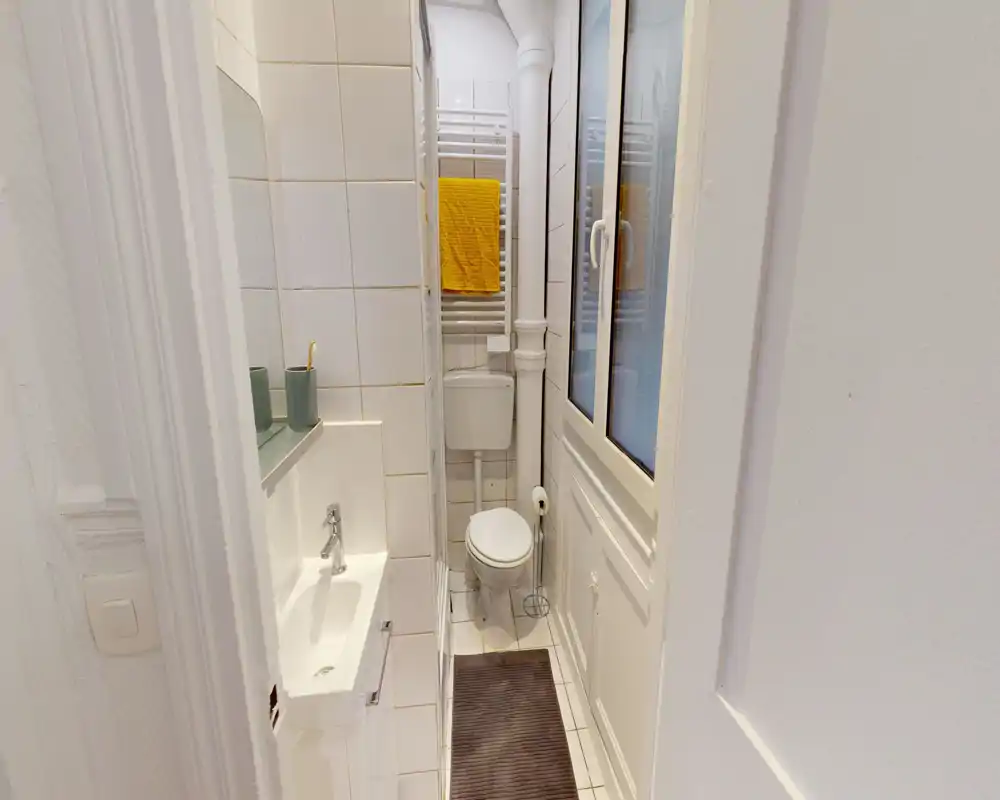 Compact, well-lit bathroom with a corner sink, wall-mounted toilet and a towel radiator. White tiles and a window create a bright, clean atmosphere ideal for small apartments.