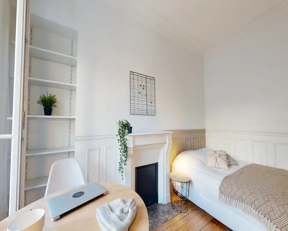 Chambre lumineuse et minimaliste avec un lit simple confortable, un sol en bois chaleureux et une décoration soignée, comprenant une petite cheminée et un bureau — idéale pour un séjour calme.