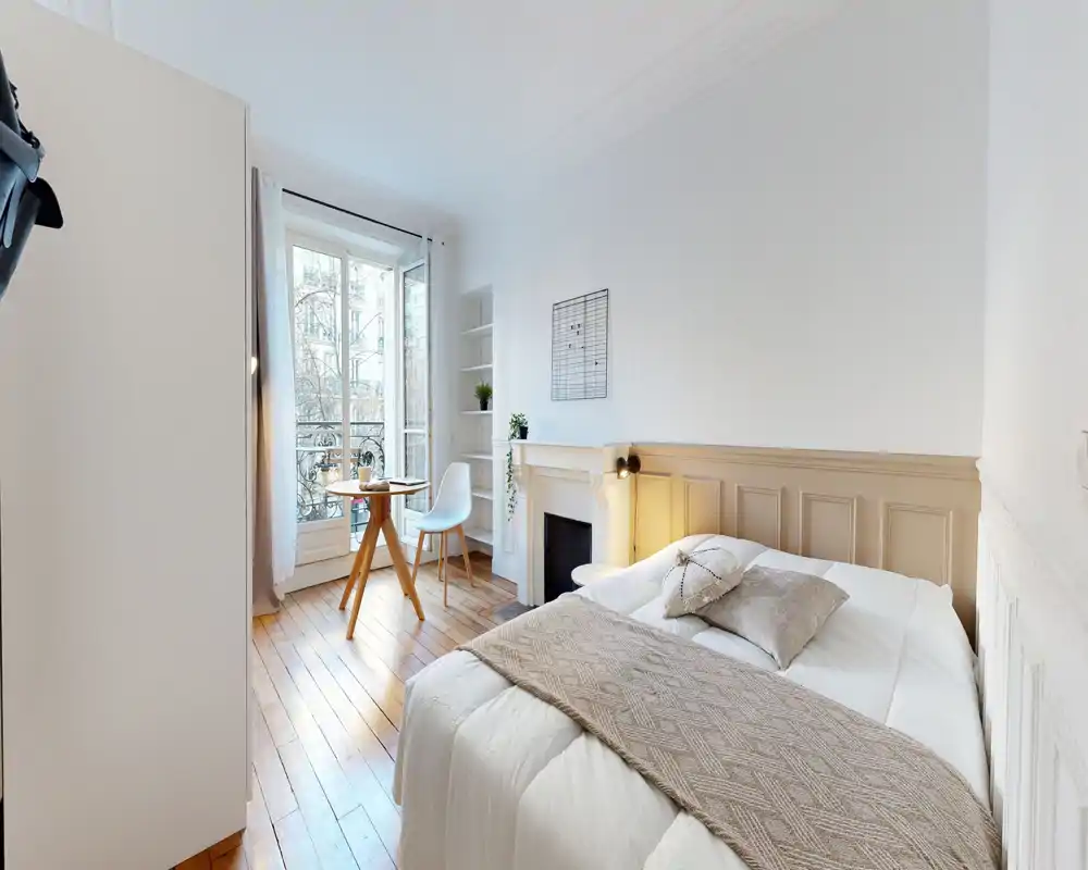Chambre lumineuse et accueillante avec un lit double bien fait, parquet chaleureux et une petite table près de la fenêtre laissant entrer une belle lumière naturelle.