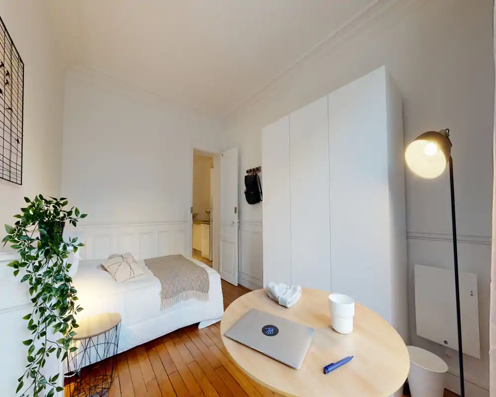 Chambre lumineuse et élégante avec lit simple, armoire et petite table de travail; parquet chaleureux et plante décorative pour une ambiance cosy.