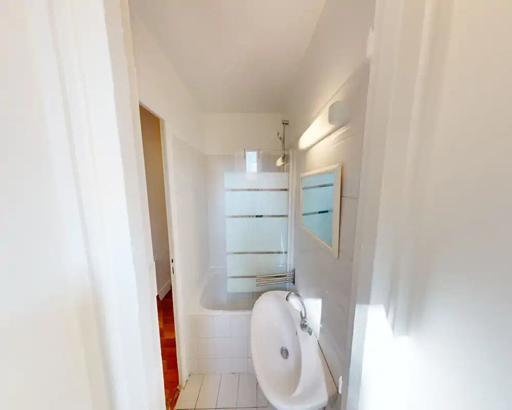 Salle de bain compacte et lumineuse avec carrelage blanc, lavabo ovale mural, baignoire avec pare-douche en verre dépoli et petit miroir — propre et fonctionnelle.
