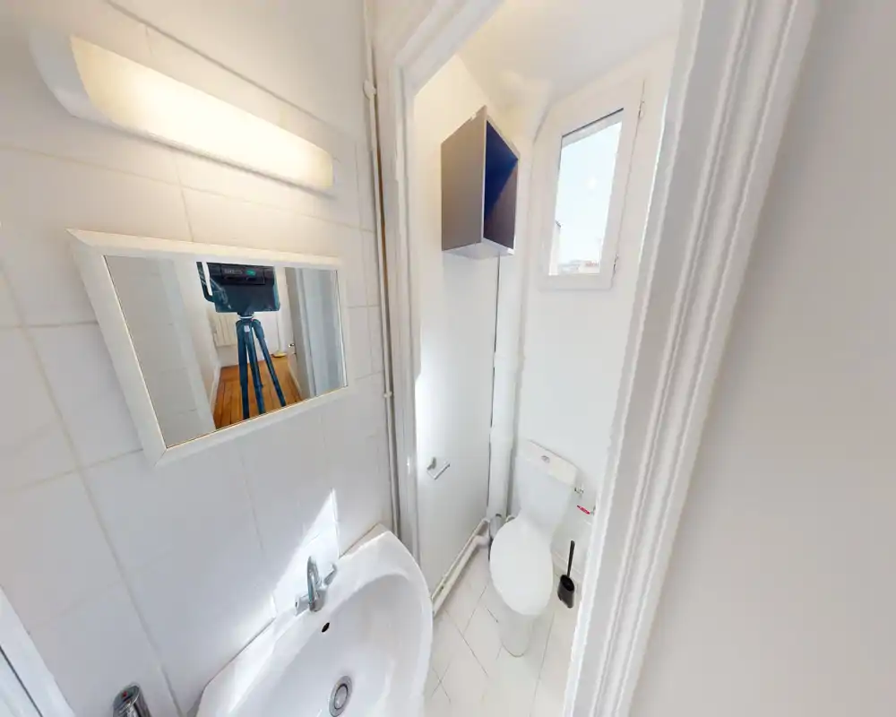 Petite salle de bain blanche avec lavabo, miroir et WC. Espace lumineux et propre, idéal pour un petit appartement.