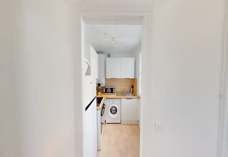 Cuisine moderne et compacte avec meubles blancs, plan de travail en bois et électroménagers intégrés (y compris lave-linge) ; espace lumineux, propre et bien organisé, parfait pour un appartement urbain.