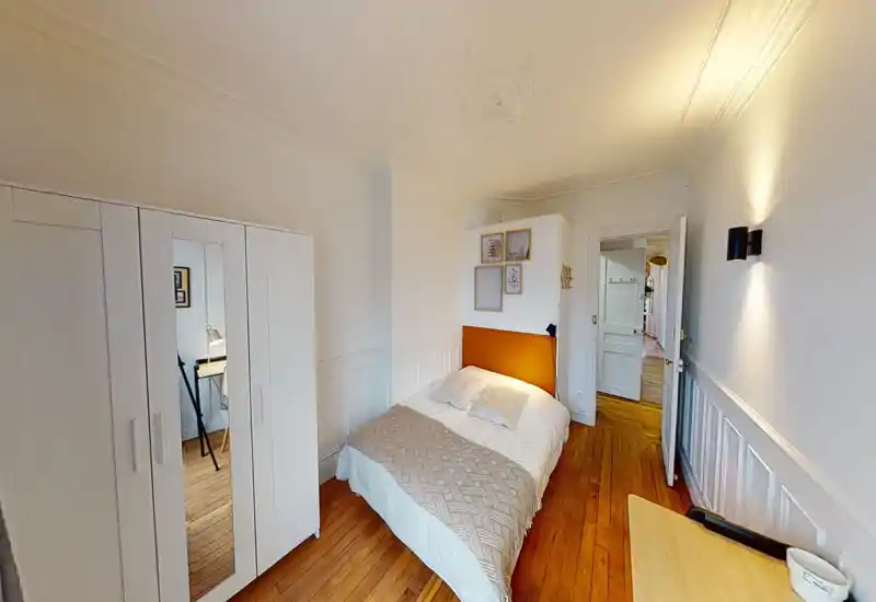 Cozy single bedroom with a warm orange headboard, neatly made bed, white wardrobe with mirrored door and wooden floors — bright, well-styled space ideal for a restful stay.