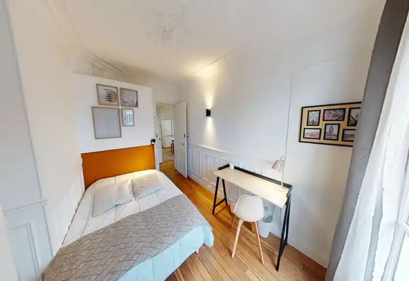 Bright, stylish bedroom with a comfortable double bed, warm orange headboard and wooden floors. A compact desk by the window provides a practical workspace; framed prints and soft textiles add character.
