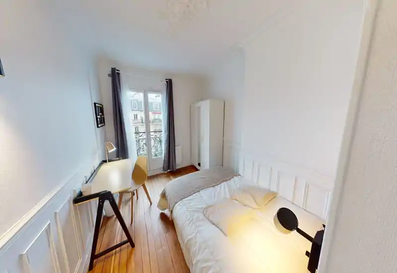 Bright, compact bedroom with a double bed, cozy throws and pillows, a small desk by the window and a freestanding wardrobe. The tall window with curtains brings natural light and overlooks a traditional Parisian-style façade.