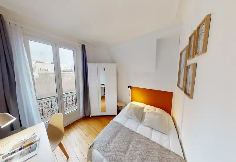 Chambre simple lumineuse et chaleureuse avec lit confortable, parquet en bois et grande fenêtre donnant sur un balcon à la parisienne. Placard blanc et bureau épurés offrent un espace fonctionnel et bien éclairé, parfait pour courts séjours.