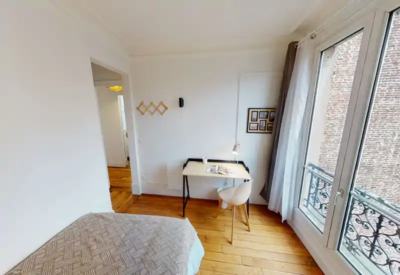 Chambre compacte et lumineuse avec un lit confortable, parquet et un petit bureau près d’une grande fenêtre offrant une belle lumière naturelle — parfaite pour une personne ou le télétravail.