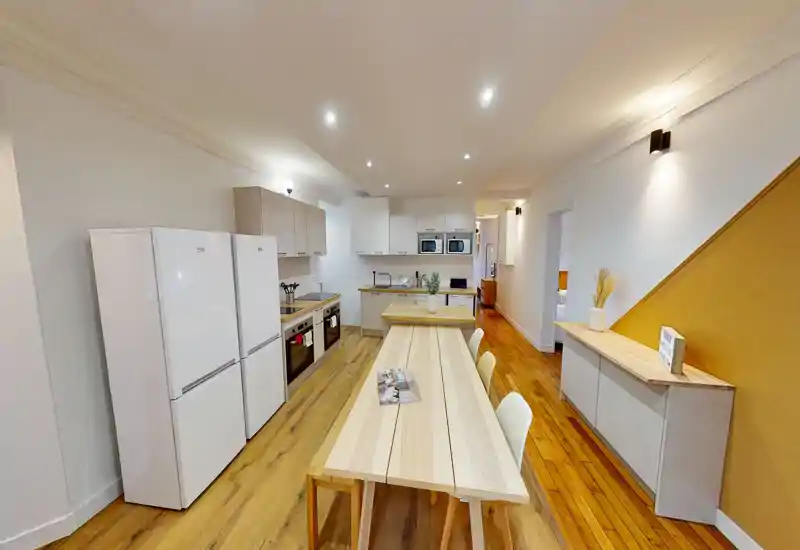 Cuisine ouverte moderne et lumineuse avec grande table en bois, double réfrigérateur, fours encastrés et meubles clairs — idéale pour cuisiner et partager des repas.