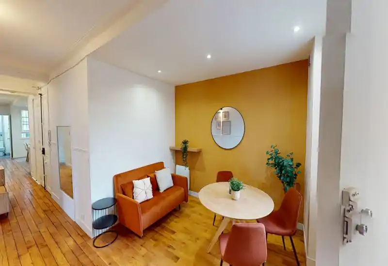 Coin salon/salle à manger chaleureux et bien mis en scène avec un petit canapé, table ronde et chaises assorties devant un mur couleur moutarde — parfait pour un séjour cosy et des photos stylées.