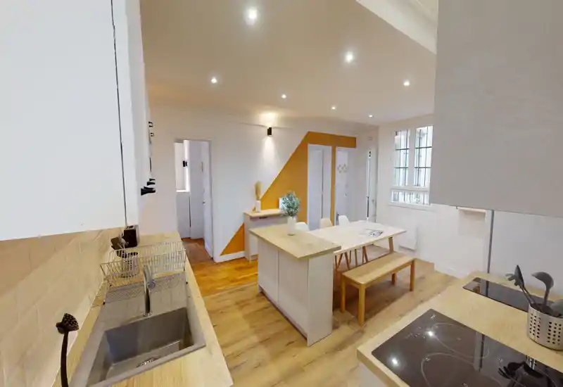 Cuisine moderne et lumineuse avec plans de travail en bois, plaque de cuisson intégrée et îlot central servant aussi d'espace repas; tons chauds et lumière naturelle pour une atmosphère accueillante.