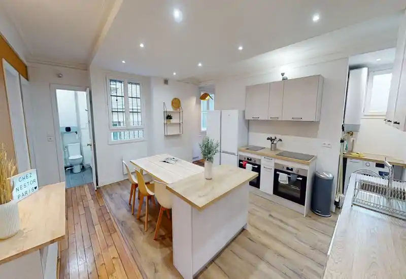 Cuisine moderne ouverte avec îlot central et table à manger, surfaces en bois clair, fours et réfrigérateur intégrés, décoration neutre et lumineuse adaptée à un appartement cosy.