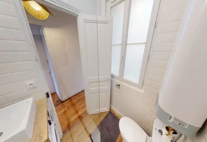 Salle de bain compacte et propre avec toilettes, lavabo et murs carrelés blancs. Une fenêtre dépoli diffuse la lumière naturelle ; le sol en bois apporte de la chaleur.