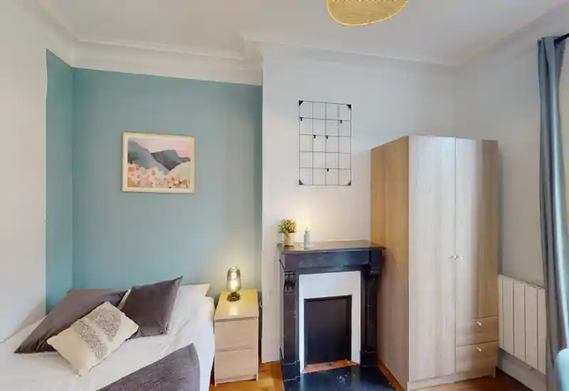 Camera da letto luminosa e ben arredata con letto singolo vicino a una parete accentata color teal, comodino con lampada, armadio e camino decorativo — perfetta per un soggiorno accogliente.