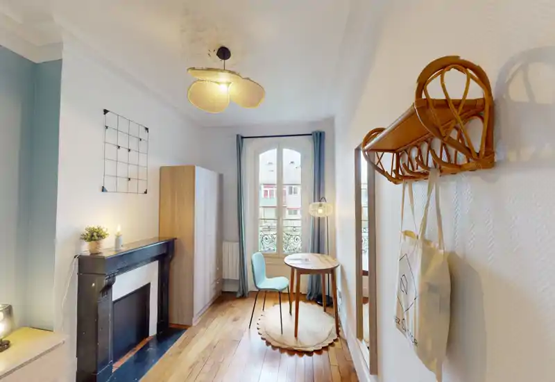 Stanza luminosa in stile camera da letto, stretta, con camino decorativo, armadio, piccolo tavolo rotondo e sedia vicino a una finestra ad arco; pavimenti in legno caldi e tessili morbidi creano un’atmosfera accogliente.
