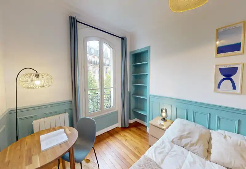 Chambre lumineuse et aménagée avec un lit confortable, parquet et grande fenêtre en arc laissant entrer la lumière naturelle. Boiseries décoratives, textiles doux et lampe de chevet créent une ambiance chaleureuse.