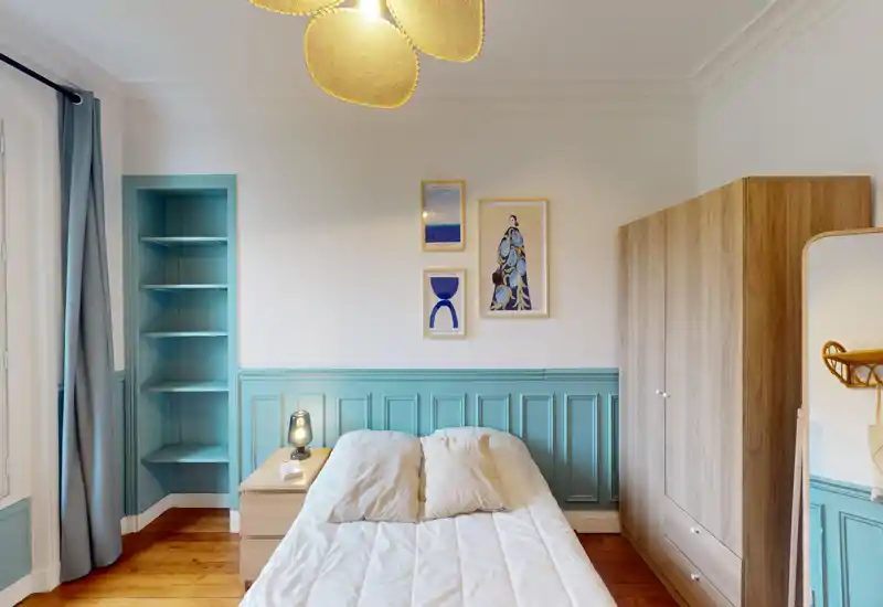 Chambre lumineuse et stylée avec lit double central, lambris bleu-vert décoratif, armoire en bois et parquet chaleureux. Linge doux et tableaux assortis créent une atmosphère accueillante, idéale pour les annonces.
