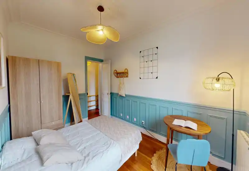 Chambre lumineuse et bien décorée avec un lit double, parquet, armoire et une petite table de lecture avec chaise — lambris bleu-vert et éclairage chaud pour une atmosphère cosy.