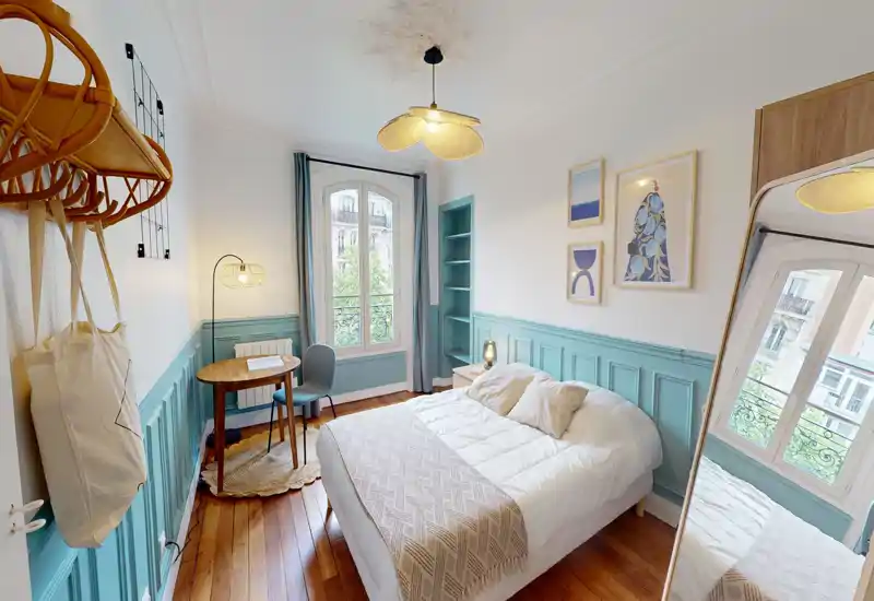 Chambre chaleureuse et stylée avec un lit double confortable, textiles doux et lumière naturelle provenant d’une grande fenêtre. Lambris turquoise, parquet en bois et petite table de travail pour un espace accueillant.