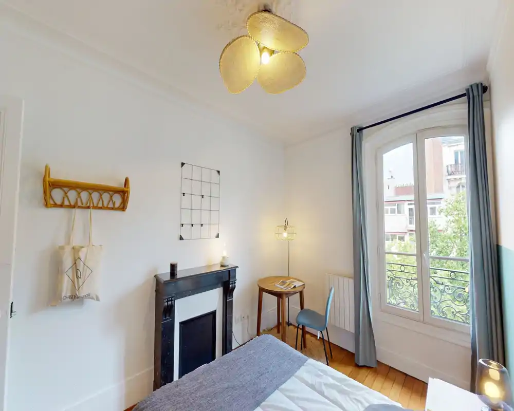 Chambre lumineuse et décorée avec lit au premier plan, petite cheminée, bureau et chaise près d'une grande fenêtre apportant une belle lumière naturelle.