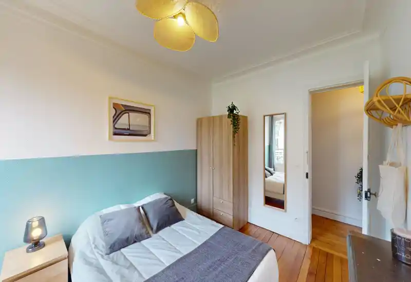 Chambre lumineuse et chaleureuse avec un lit double bien fait, textiles confortables et mur d'accent vert-bleu. Sol en bois chaleureux, grande armoire, table de chevet avec lampe et suspension décorative.