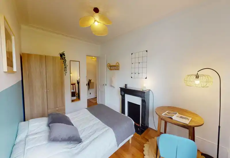 Chambre lumineuse et stylée avec lit double confortable, armoire en bois, cheminée décorative et un petit espace de travail. Lumières chaudes et textiles doux créent une atmosphère accueillante, idéale pour une annonce.