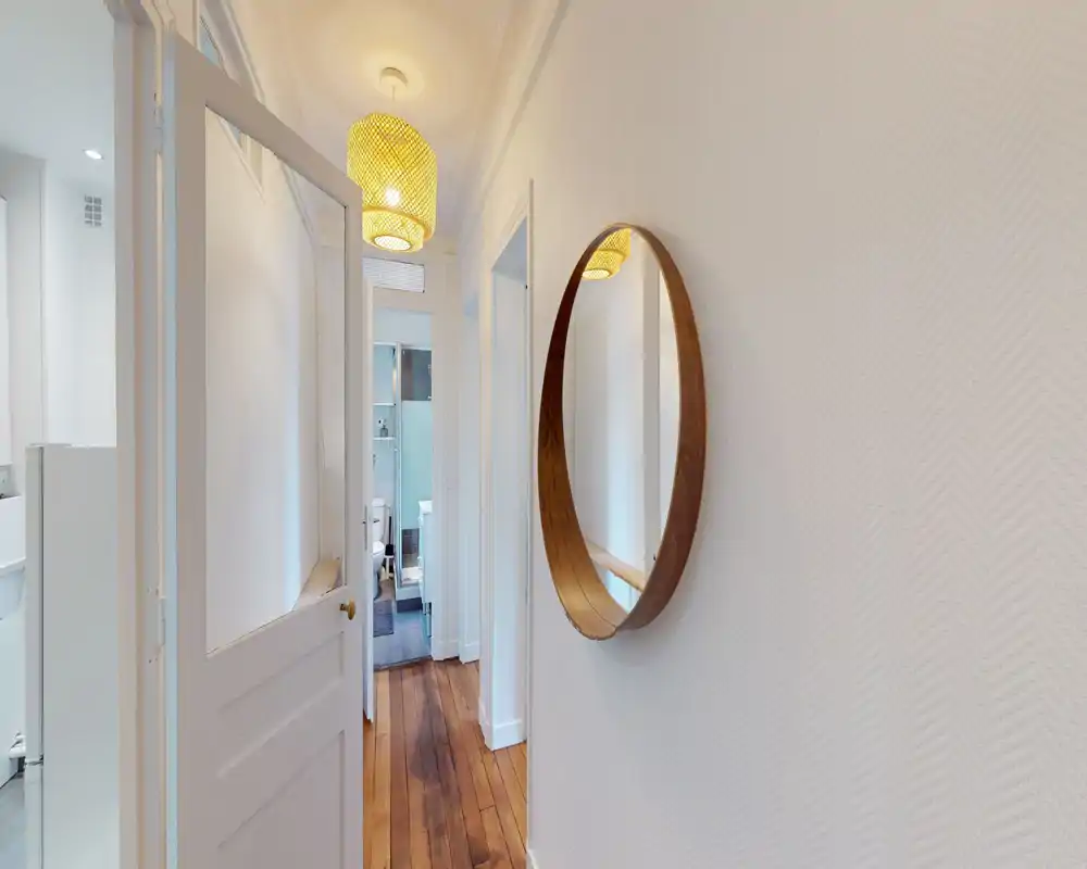 Entrée lumineuse et étroite avec parquet, miroir mural rond et suspension chaleureuse — espace d’accueil propre et accueillant.