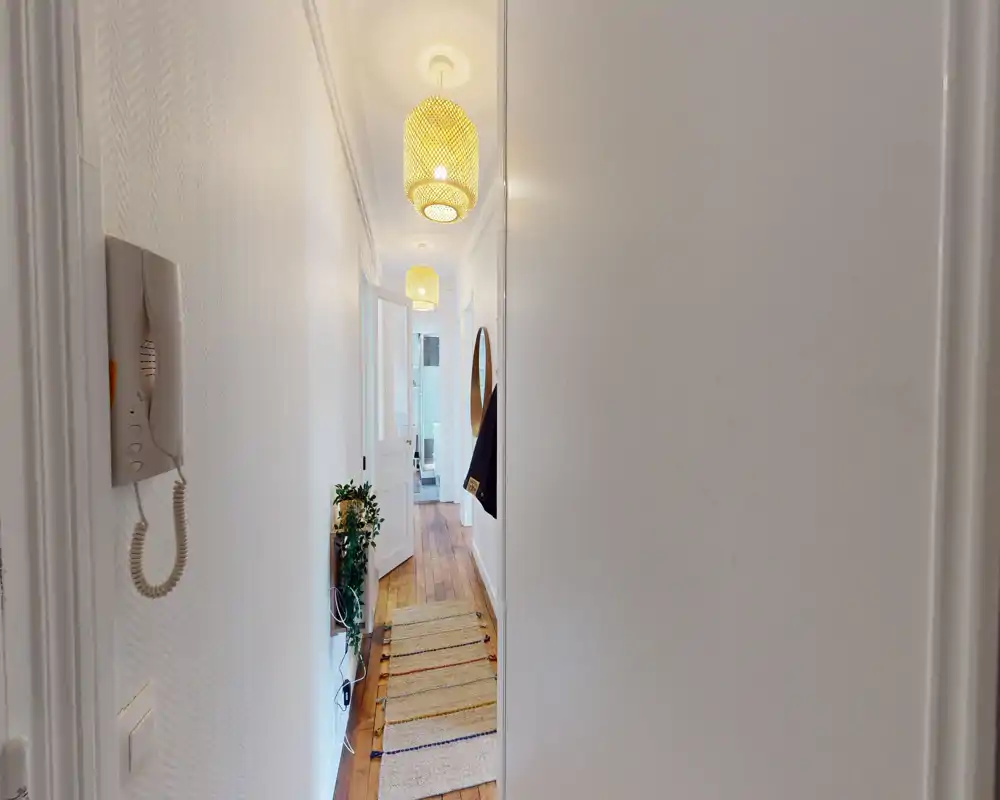 Entrée lumineuse et étroite avec sol en bois, suspensions et un tapis couloir menant au reste de l'appartement.
