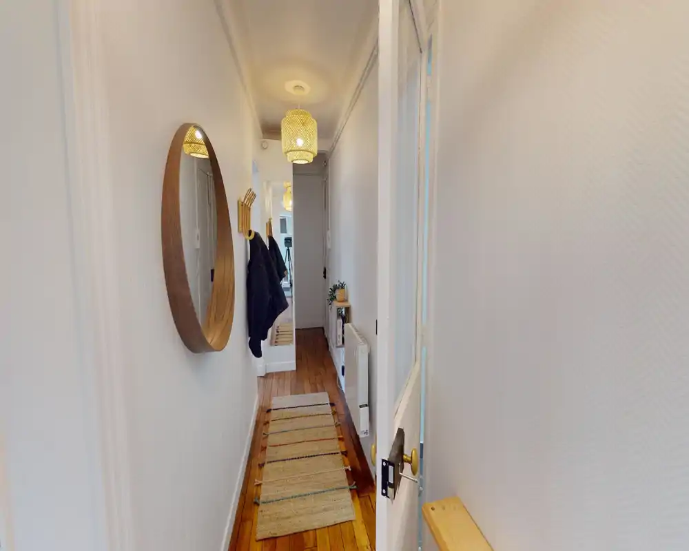 Entrée/couloir lumineux et étroit avec sol en bois, grand miroir mural rond, patères et un tapis coureur — suspension chaude qui apporte une ambiance accueillante.