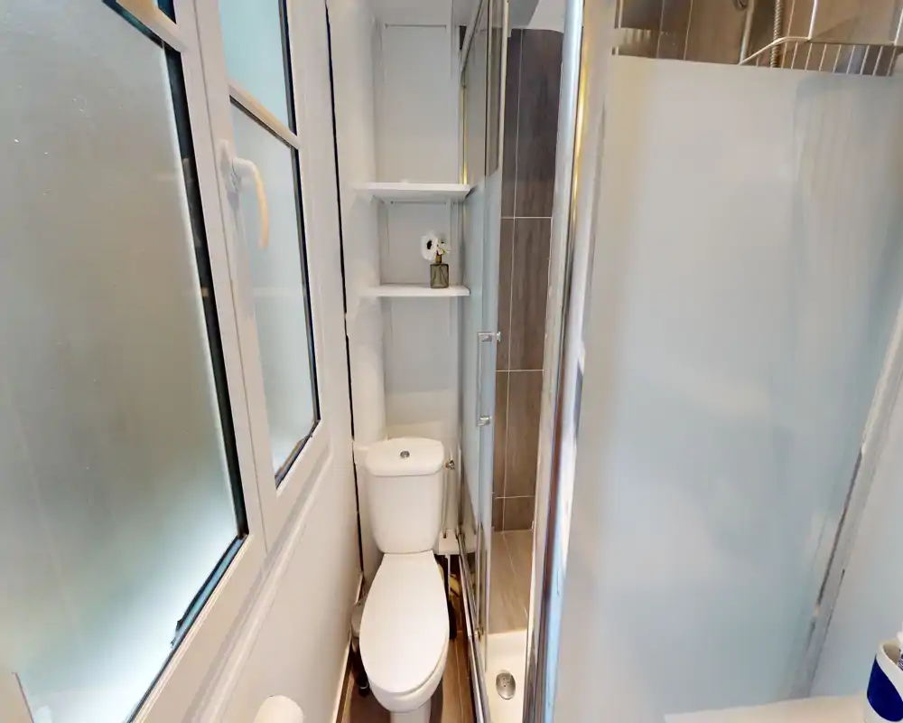 Salle de bains compacte et lumineuse avec WC, cabine de douche d'angle et étagères à côté du réservoir. Finitions modernes et fenêtre dépoli pour l'intimité.