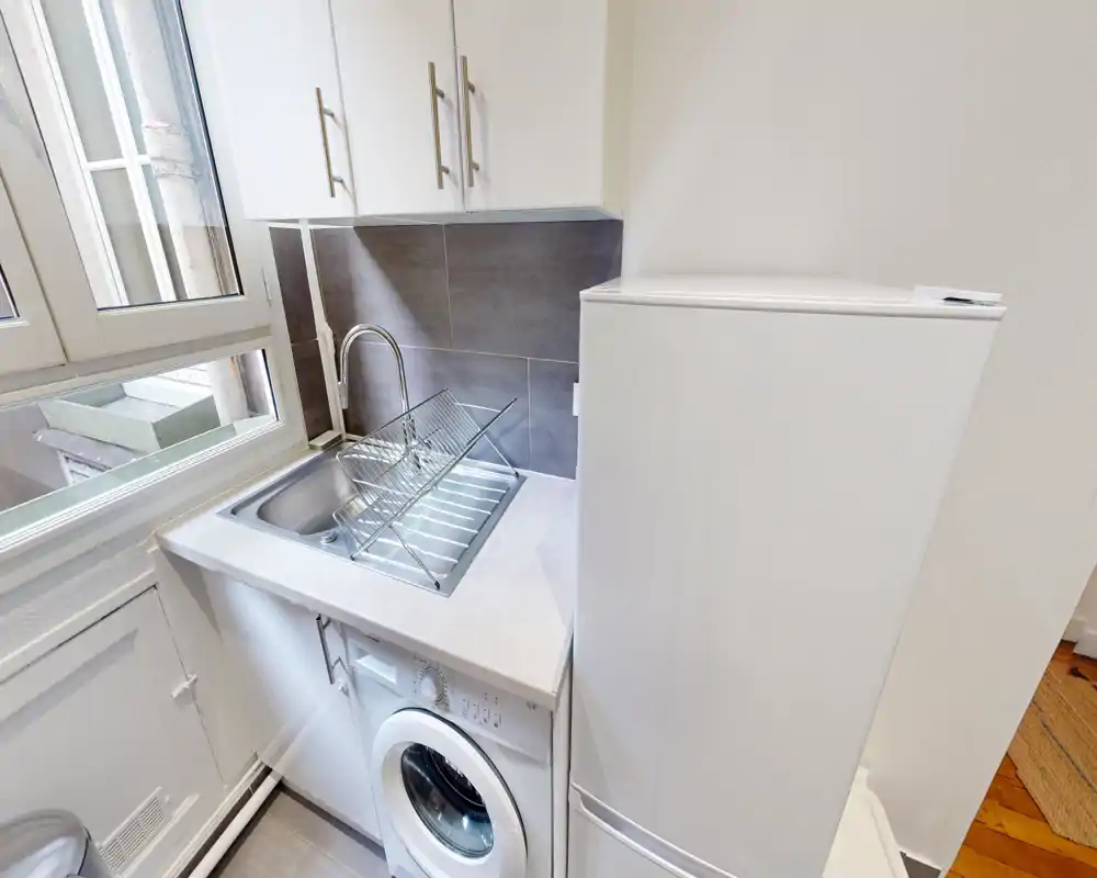 Kitchenette compacte avec évier en inox, égouttoir, lave-linge sous-plan et grand réfrigérateur. Lumière naturelle par la fenêtre et carrelage neutre créent un espace propre et fonctionnel, parfait pour un petit appartement.