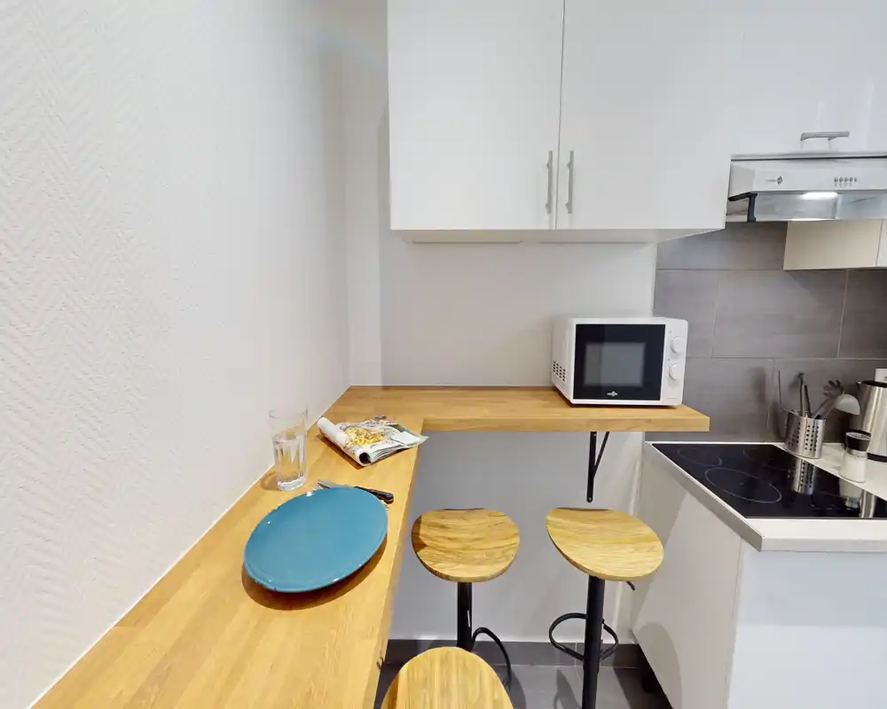 Kitchenette compacte et moderne avec plan de travail en bois et trois tabourets, plaque de cuisson électrique et micro-ondes — pratique et chaleureuse pour un petit appartement.