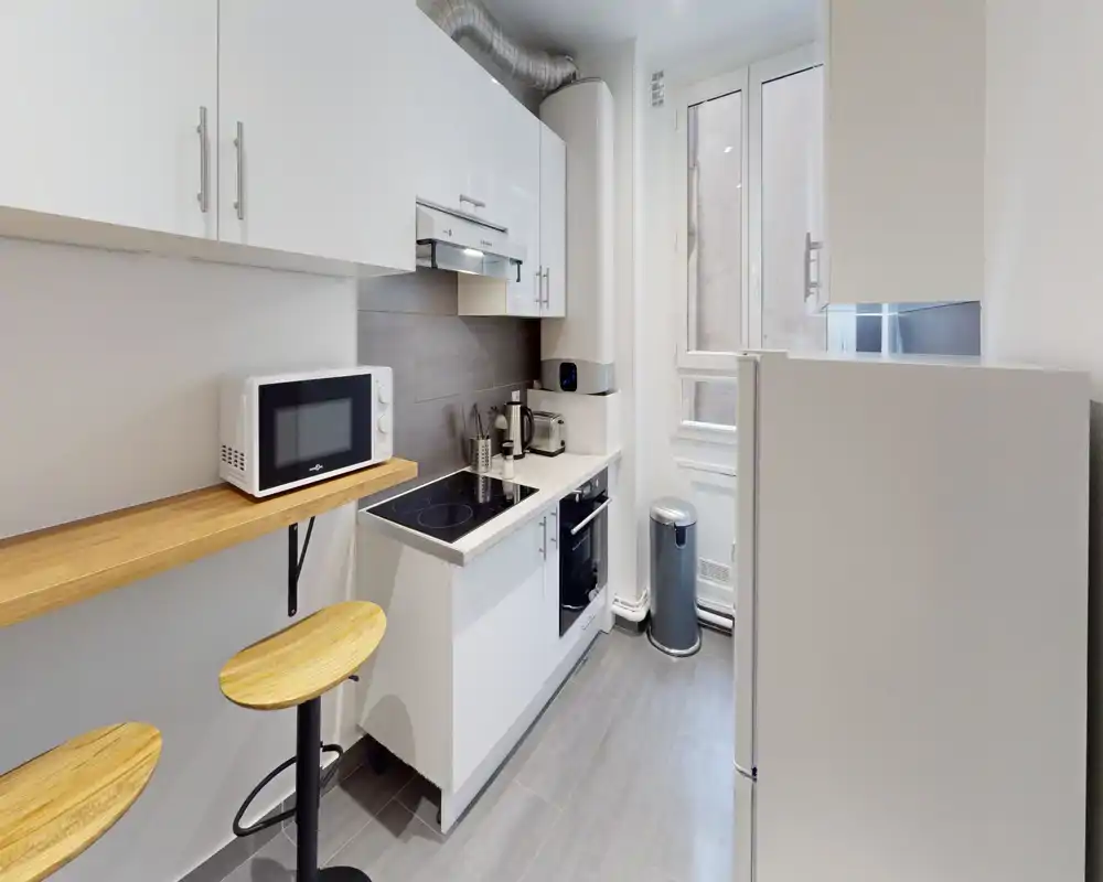 Cuisine moderne compacte avec placards blancs, plaque à induction, micro-ondes et bar avec tabourets en bois — lumineuse et aménagée de façon efficace.