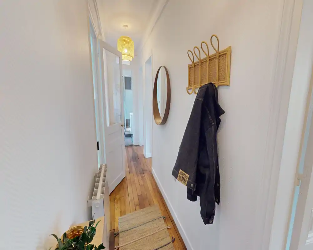 Couloir d'entrée lumineux et étroit avec sol en bois, patères murales et miroir rond — passage soigné et accueillant vers le reste du logement.