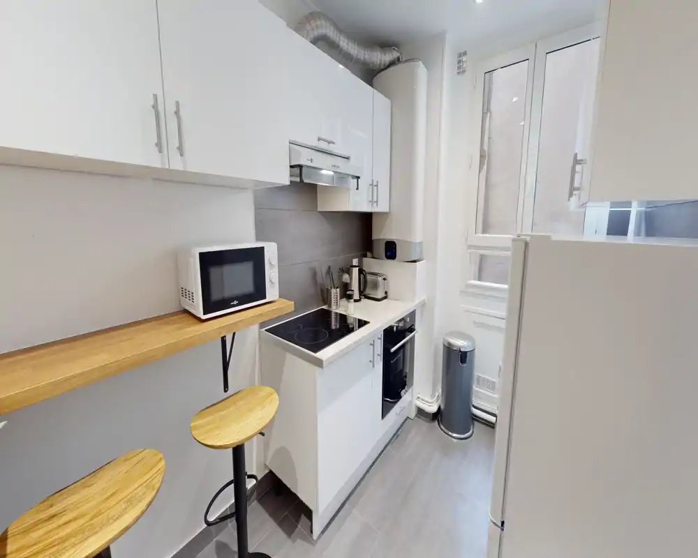 Cuisine compacte et moderne avec placards blancs, plaque de cuisson électrique, four intégré et petit bar avec tabourets en bois. Une fenêtre apporte une bonne lumière naturelle, agencement optimisé pour un appartement urbain.