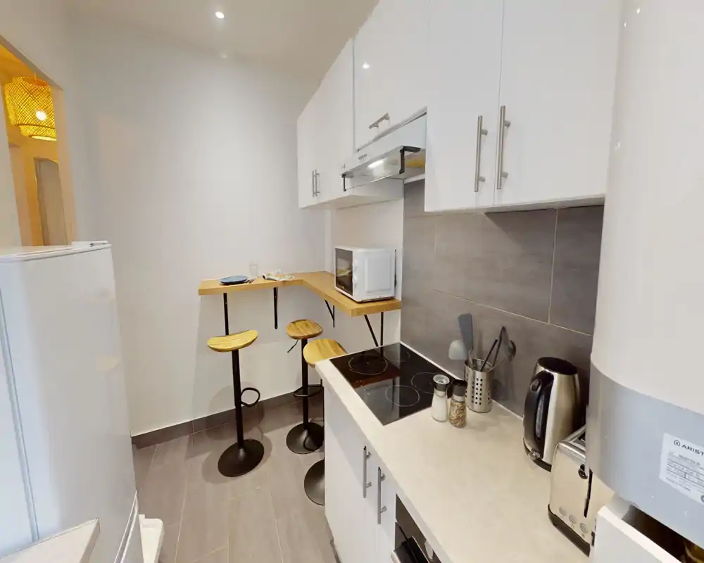 Cuisine moderne et compacte avec meubles blancs, plaque à induction, bouilloire et petit bar en bois avec tabourets — lumineuse et bien agencée.
