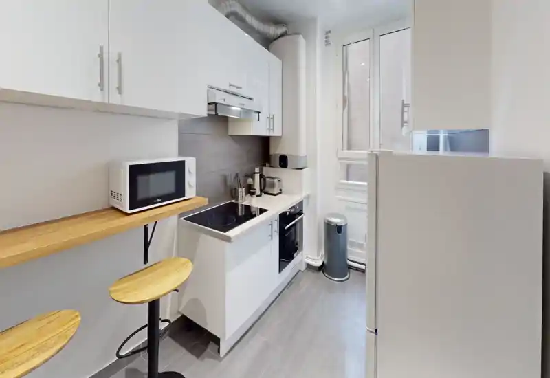 Cuisine moderne compacte avec placards blancs, plaque à induction, micro-ondes et bar avec tabourets en bois — lumineuse et aménagée de façon efficace.