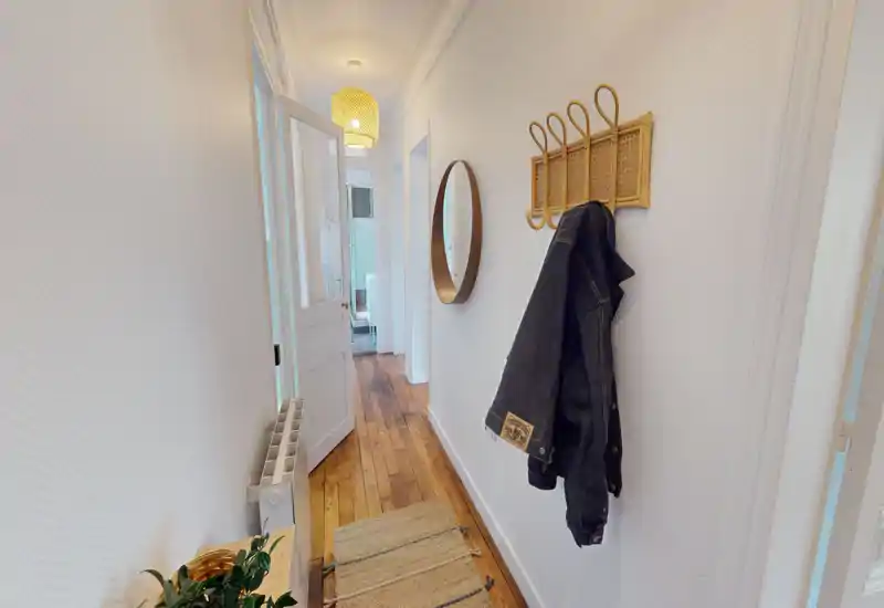 Couloir d'entrée lumineux et étroit avec sol en bois, patères murales et miroir rond — passage soigné et accueillant vers le reste du logement.