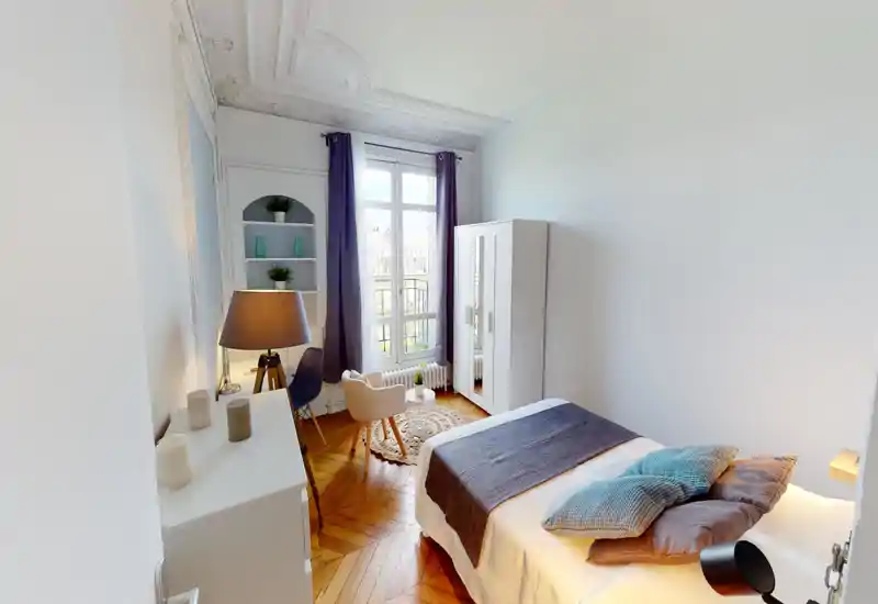 Chambre lumineuse et élégante avec un lit double confortable, coussins décoratifs et plaid. Sol en parquet, armoire, coin lecture près d'une grande fenêtre et moulures au plafond.