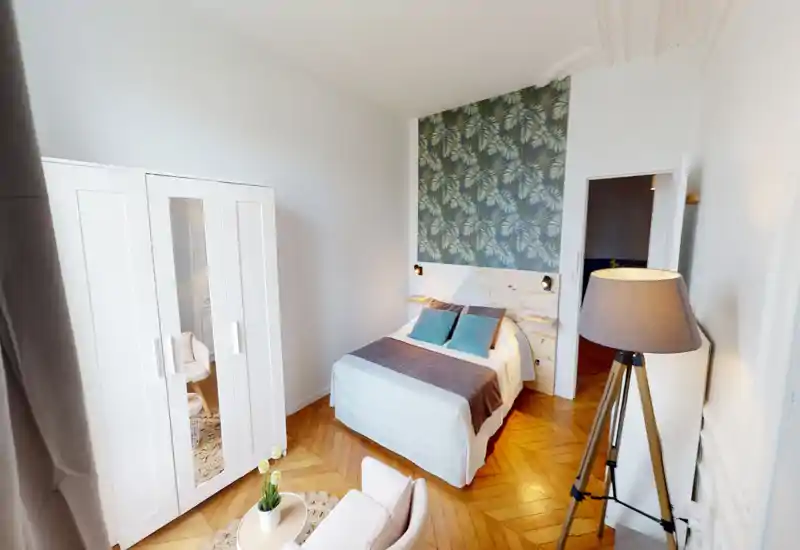 Chambre lumineuse et élégante avec un lit double devant un panneau de papier peint feuillu ; parquet chaleureux, lampes de chevet, armoire avec miroir et lampadaire complètent cet espace cosy et bien mis en scène.