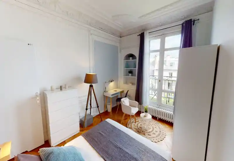 Chambre lumineuse et élégante avec un grand lit au premier plan, moulures décoratives, un coin lecture près de la fenêtre et une belle lumière naturelle.
