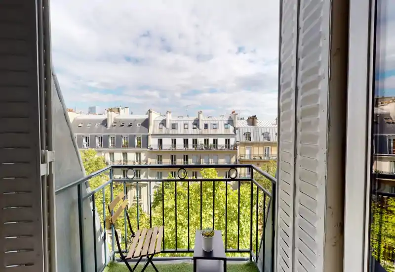 Petit balcon ensoleillé avec chaise pliante, petite table et plante en pot, offrant une vue sur les toits parisiens et une rue bordée d'arbres.