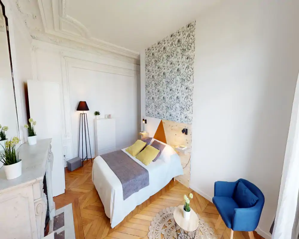 Chambre lumineuse et élégante avec un lit double centré devant un mur à motif, parquet chaleureux et touches décoratives soignées comme un fauteuil bleu et des lampes de chevet.