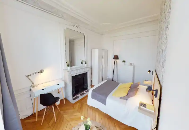 Chambre lumineuse et élégante avec un grand lit confortable, moulures décoratives, parquet en bois et un petit bureau — idéale pour un séjour cosy.