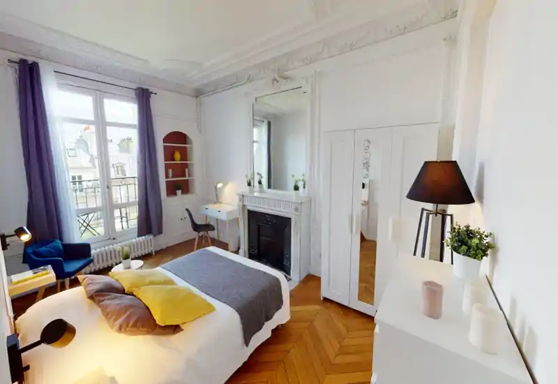 Chambre lumineuse et élégante avec grand lit, coussins décoratifs et une cheminée classique. La pièce dispose de grandes fenêtres avec rideaux violets, un fauteuil cosy, un petit bureau et un parquet—idéal pour un séjour confortable dans un appartement raffiné.