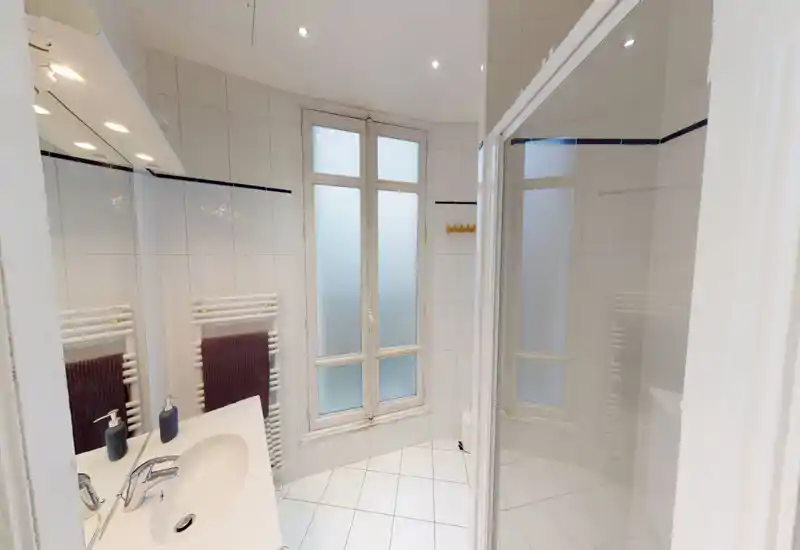 Salle de bain moderne et lumineuse avec carrelage blanc, lavabo et miroir, sèche-serviettes et cabine de douche en verre; fenêtres dépoli pour intimité et lumière naturelle.