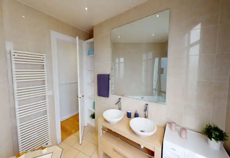 Salle de bains moderne et lumineuse avec deux vasques, grand miroir, murs carrelés et sèche-serviettes. Agencement propre et bien éclairé, idéal pour une annonce.